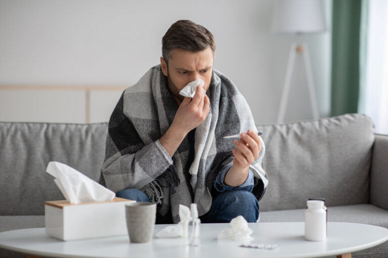 Picture of a sick man sitting on a couch under a blanket as he holds a tissue to his nose and looks at a thermometer.