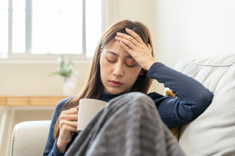 Picture of a sick woman sitting on a couch under a blanket, holding a mug and touching her forehead with the other hand.