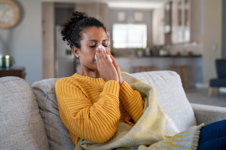 Picture of a sick woman sitting on a couch under a blanket, blowing her nose into a tissue.