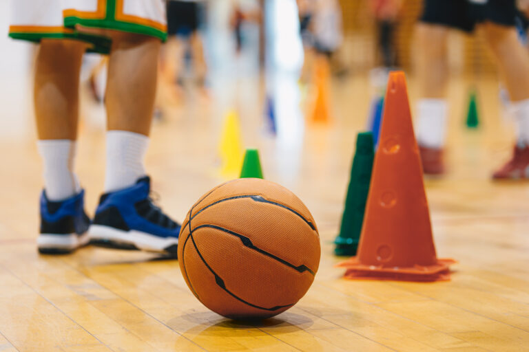 Picture of a basketball and safety cones on a gymnasium floor with children playing in the background.