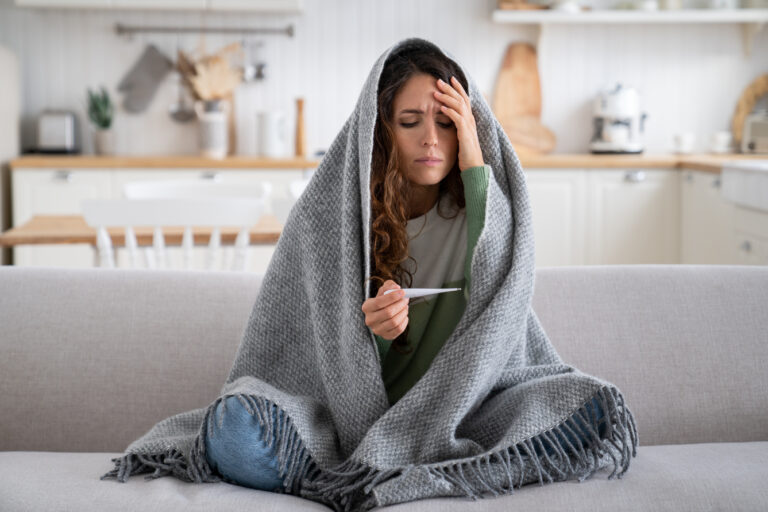 Picture of a sick woman sitting on a couch, wrapped in a blanket, feeling her forehead and holding a thermometer.