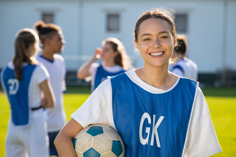 Picture of a teenage girl posing with a soccer ball in front of other soccer players.