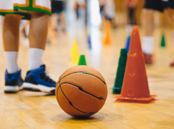 Children Practicing Basketball on School Court. Basketball Training Game Background. Basketball and Training Cones on Wooden Floor Close Up with Blurred Players Playing Basketball Game in Background