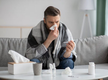 Bearded man covered in blanket feeling sick, checking for fever with thermometer