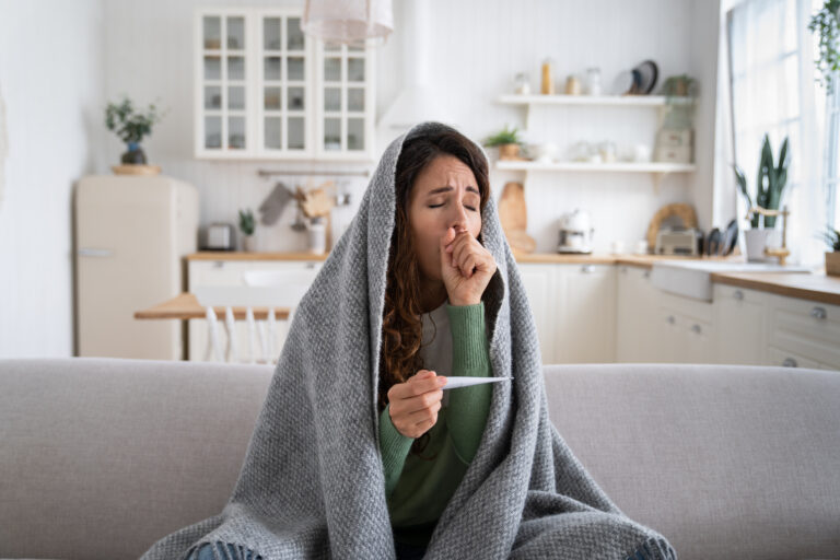Picture of a sick woman holding a thermometer and sitting on a couch wrapped in a blanket.