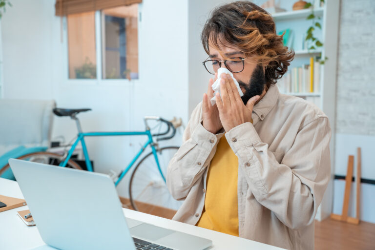 Picture of a sick man blowing his nose into a tissue while he sits in front of a laptop.