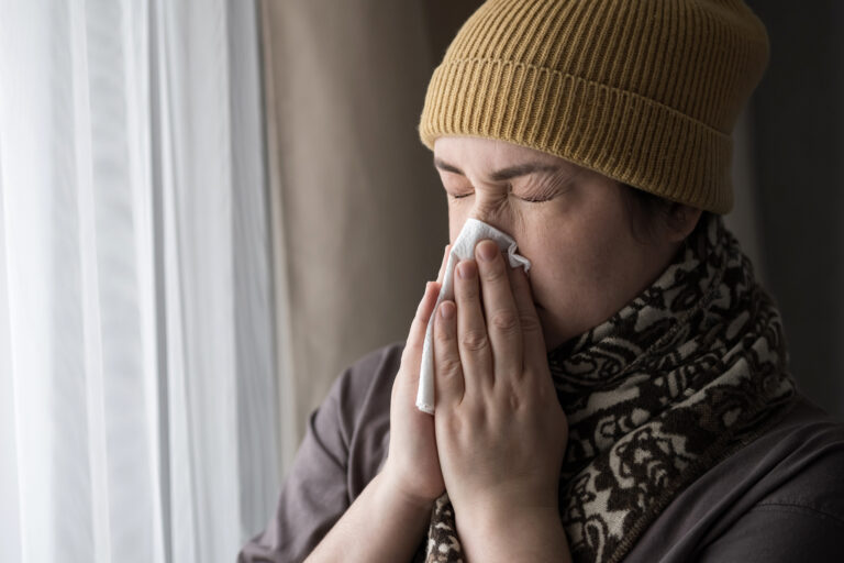 Picture of a sick woman blowing her nose into a tissue while standing in front of a window.