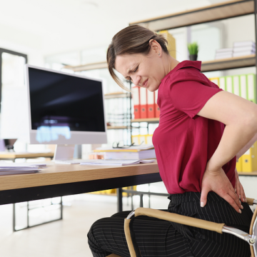 A woman holds her lower back in pain while sitting in an office chair