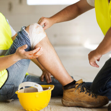 Picture of an injured construction worker sitting down with a bandage on his knee and being assisted by a coworker.