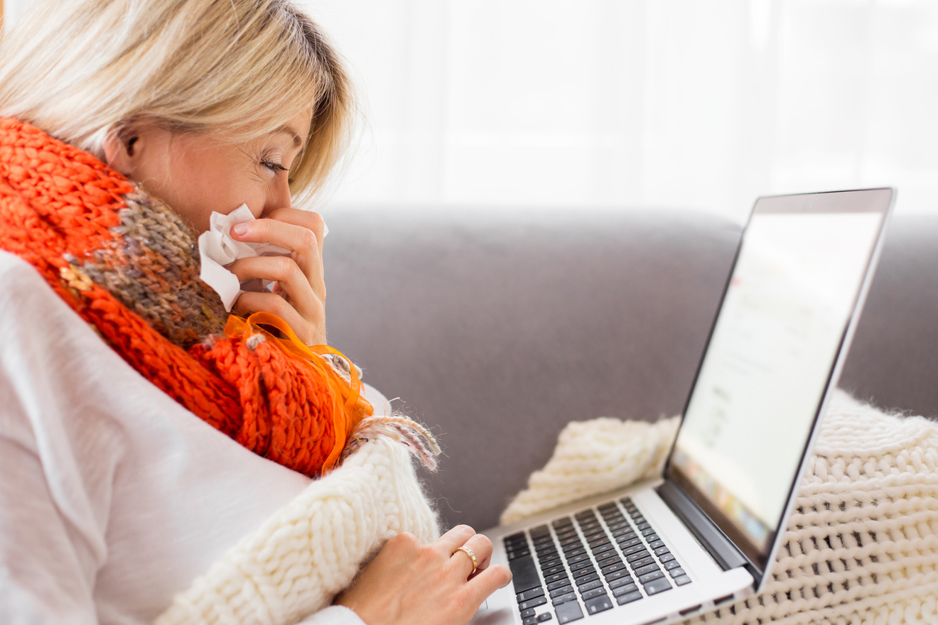 Picture of a woman sitting on a couch and blowing her nose into a tissue during a telehealth appointment.