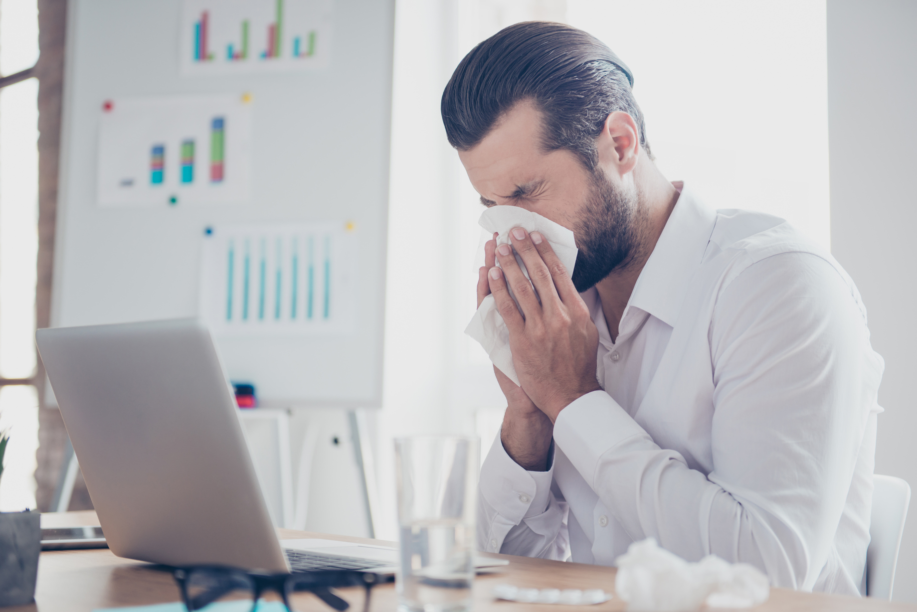 Picture of a man sneezing into a tissue while sitting in front of a laptop.
